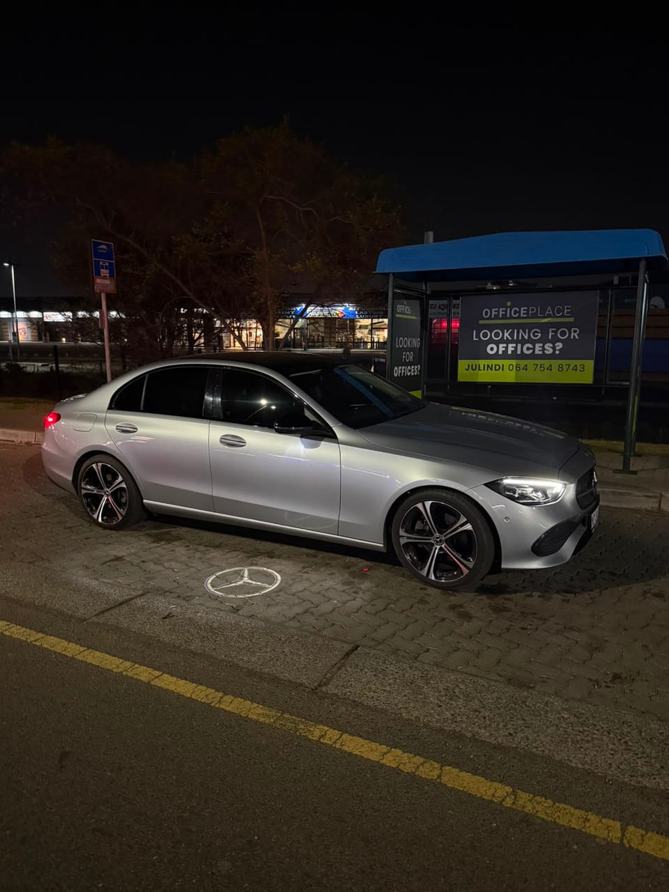 A silver Mercedes-Benz waiting at the kerb at night, its badge projected onto the pavement